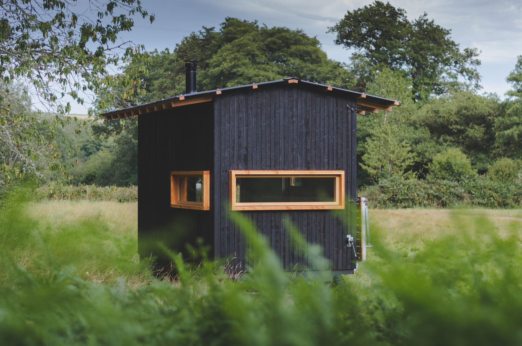 off grid tiny wooden nomad cabin with black cladding in a sunny field